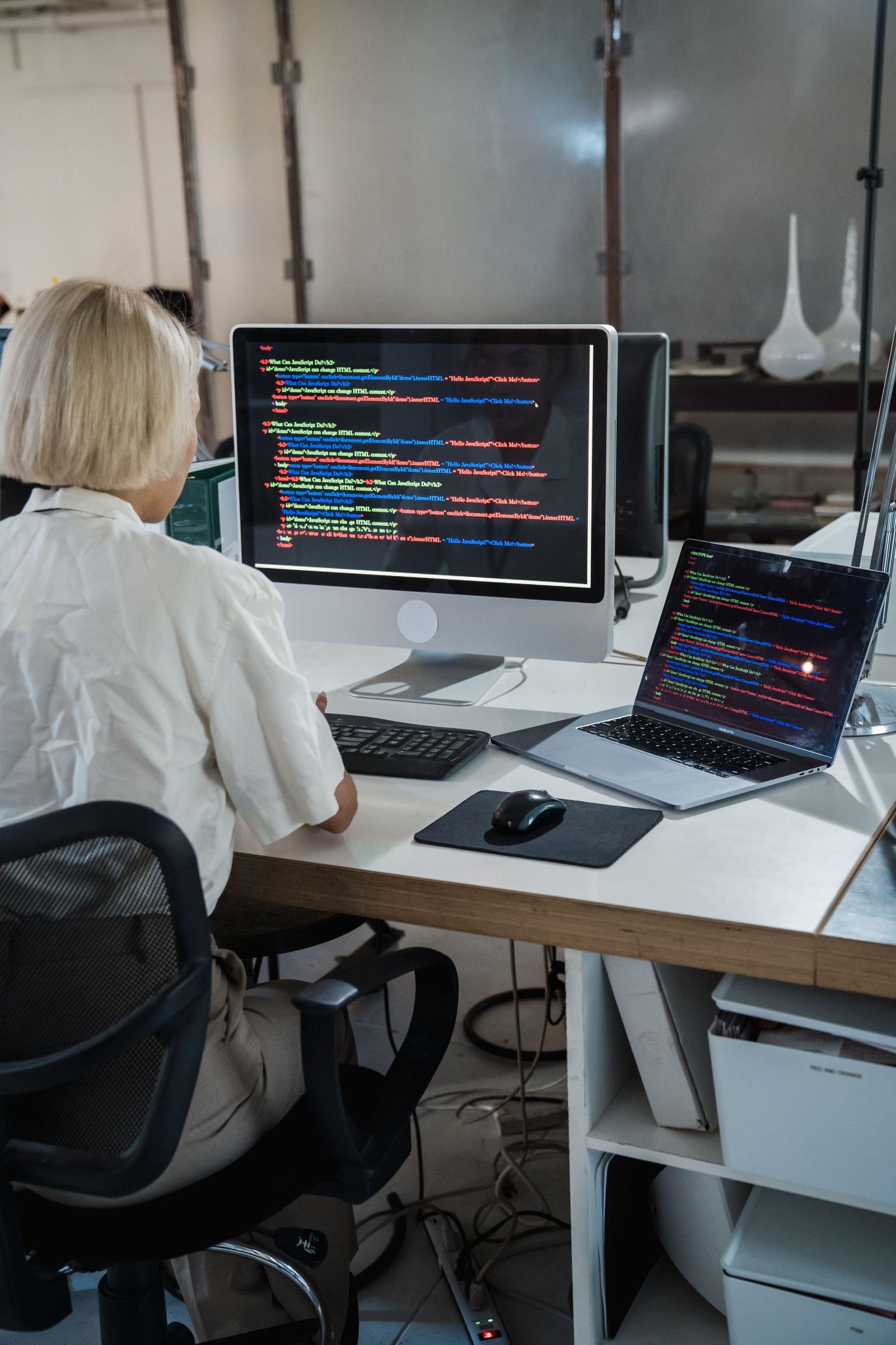 Mulher de cabelo curto loiro vista de costas, trabalhando em uma mesa com um monitor e um notebook, ambos exibindo código.