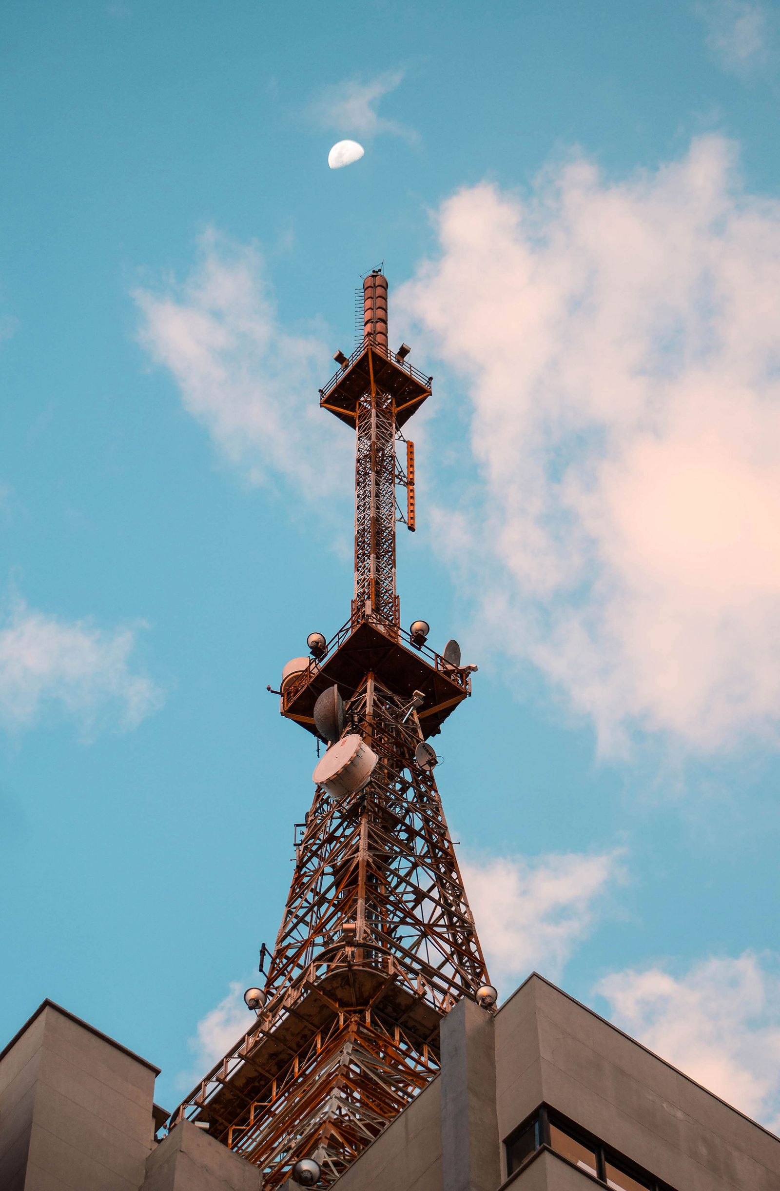 Torre de telecomunicações no alto de um edifício, com a lua em um céu azul.
