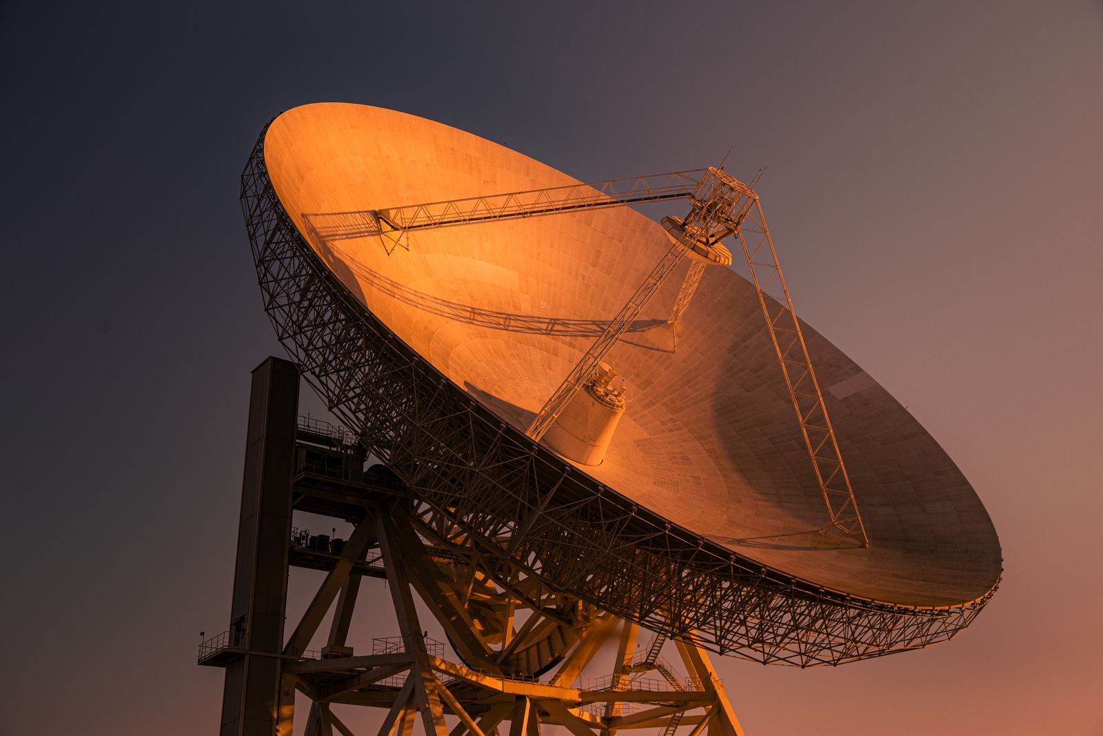 Grande antena parabólica de rádio iluminada em laranja ao anoitecer.