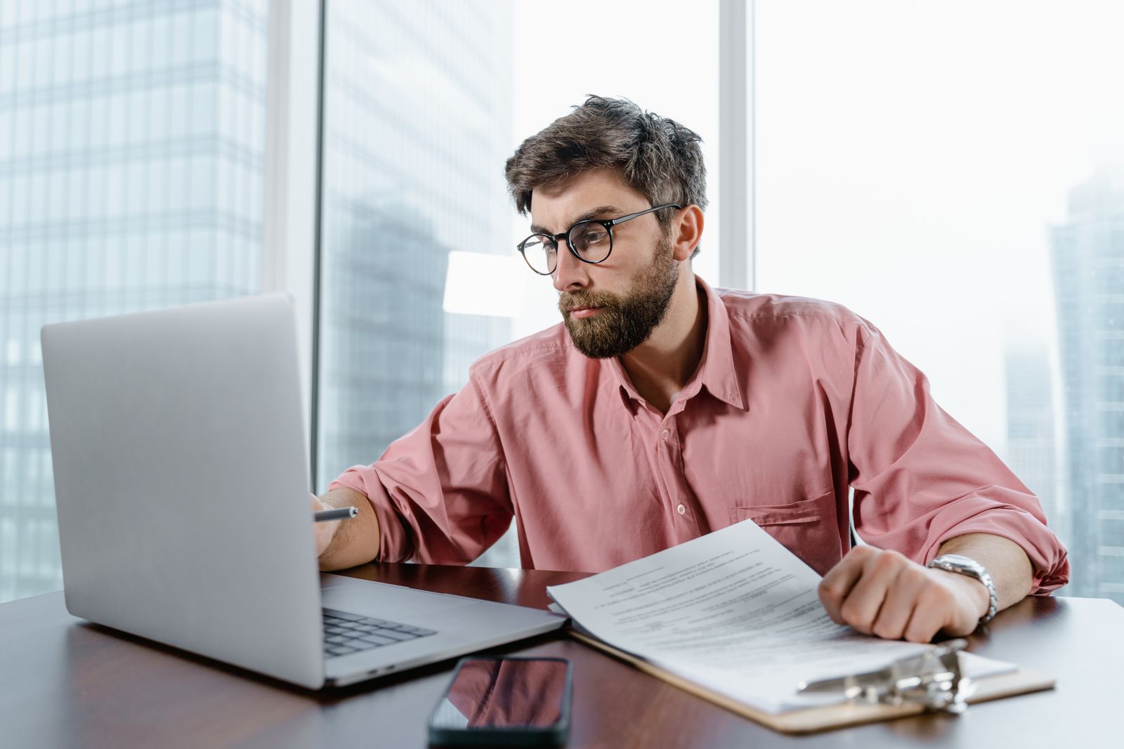 Homem barbudo de camisa rosa trabalhando em um notebook em uma mesa com papéis, com o horizonte da cidade na janela.